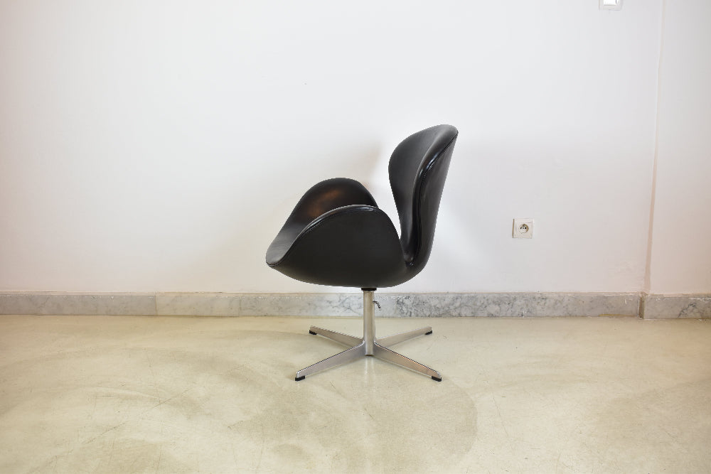 A modern black leather swivel chair with a curved shell design and a chrome four-star base, set against a plain white wall and light beige floor.